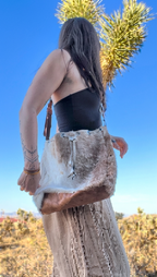 A woman outdoors wearing a black strapless top and a long beige fringe skirt, carrying a large natural fur shoulder bag with brown leather trim. The background shows a bright blue sky and a Joshua tree.