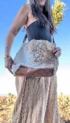 A woman outdoors wearing a black strapless top and a beige maxi skirt, holding a large shoulder bag made of natural animal fur with a brown leather base and silver-toned hardware.