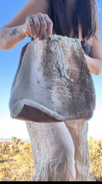 A person outdoors holding a large handbag made from animal hide, with a brown and cream mottled pattern and a silver-toned cross charm hanging from the top.