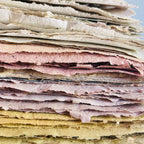 A close-up stack of textured, handmade paper sheets in various colors including cream, pink, purple, and yellow, stacked on a wooden surface.