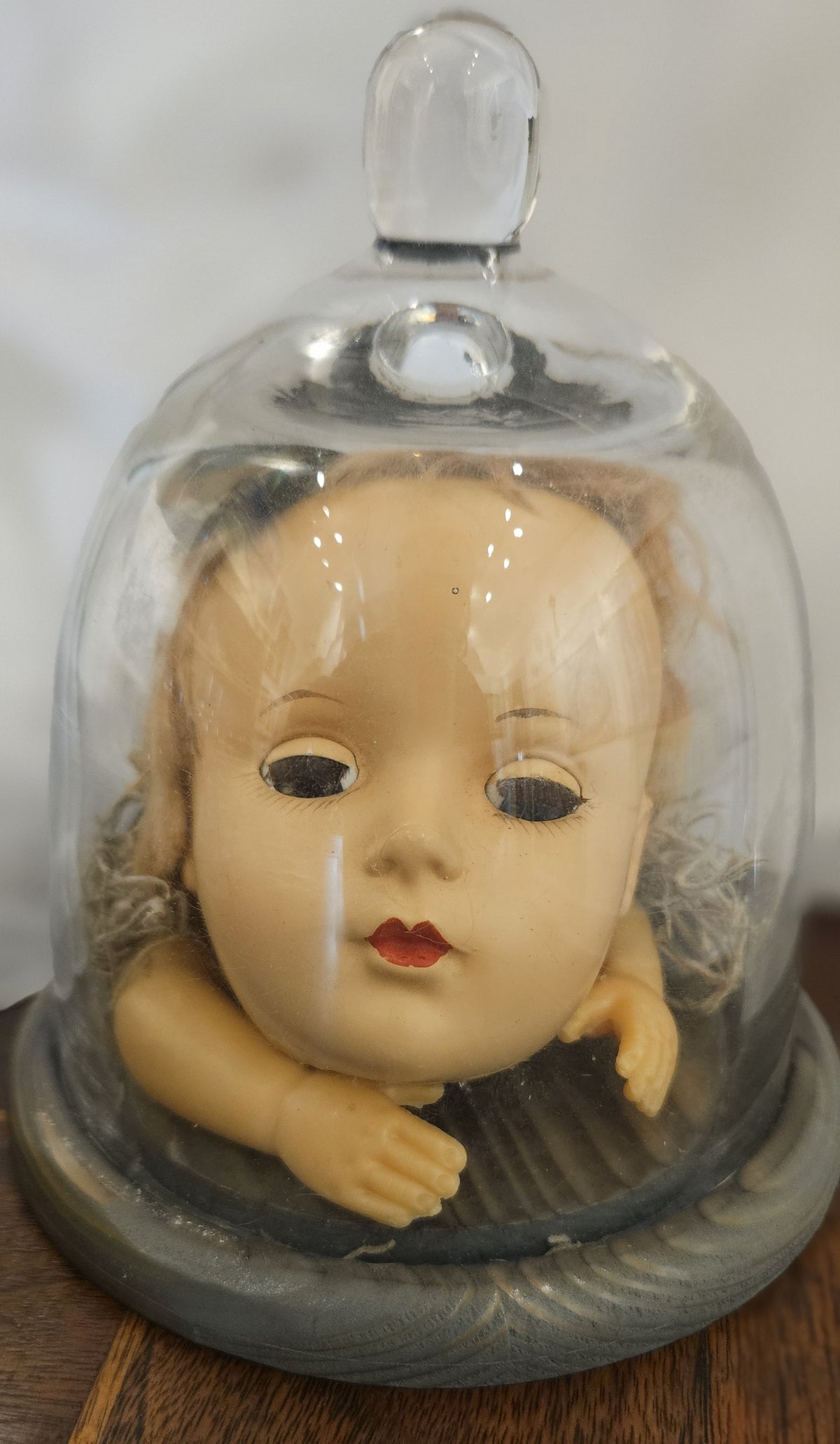 alt="Vintage doll head with hands nestled under a clear glass cloche bell jar on a carved round pedestal base"