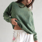 Person wearing a green sweater and white pants with floral patterns on a plain background