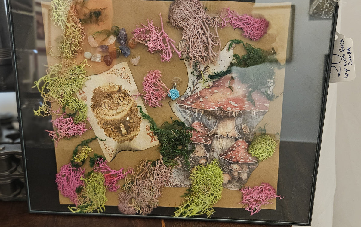 Handcrafted framed mixed-media wall art featuring an Alice in Wonderland Cheshire Cat playing card, red toadstool illustrations, colorful preserved moss, and crystal chips in a black frame.