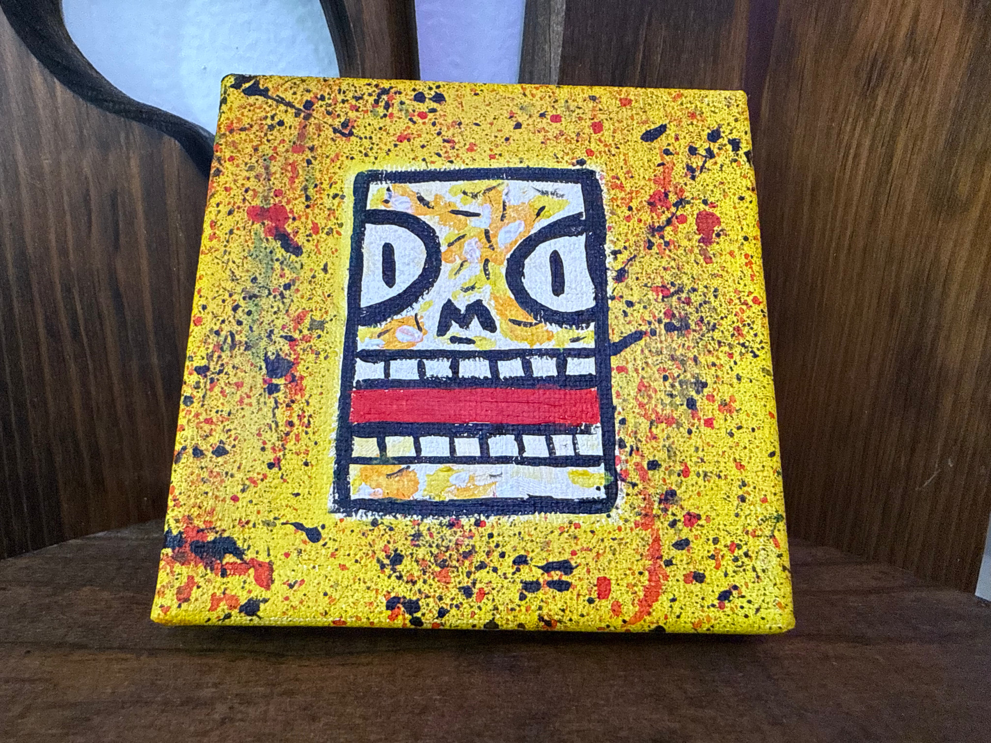"Anger" by Shelf4Life 4x4 mini canvas painting featuring a stylized, angry square face with wide eyes and gritted teeth on a textured yellow background with red and black paint splatters, displayed on a wooden chair.