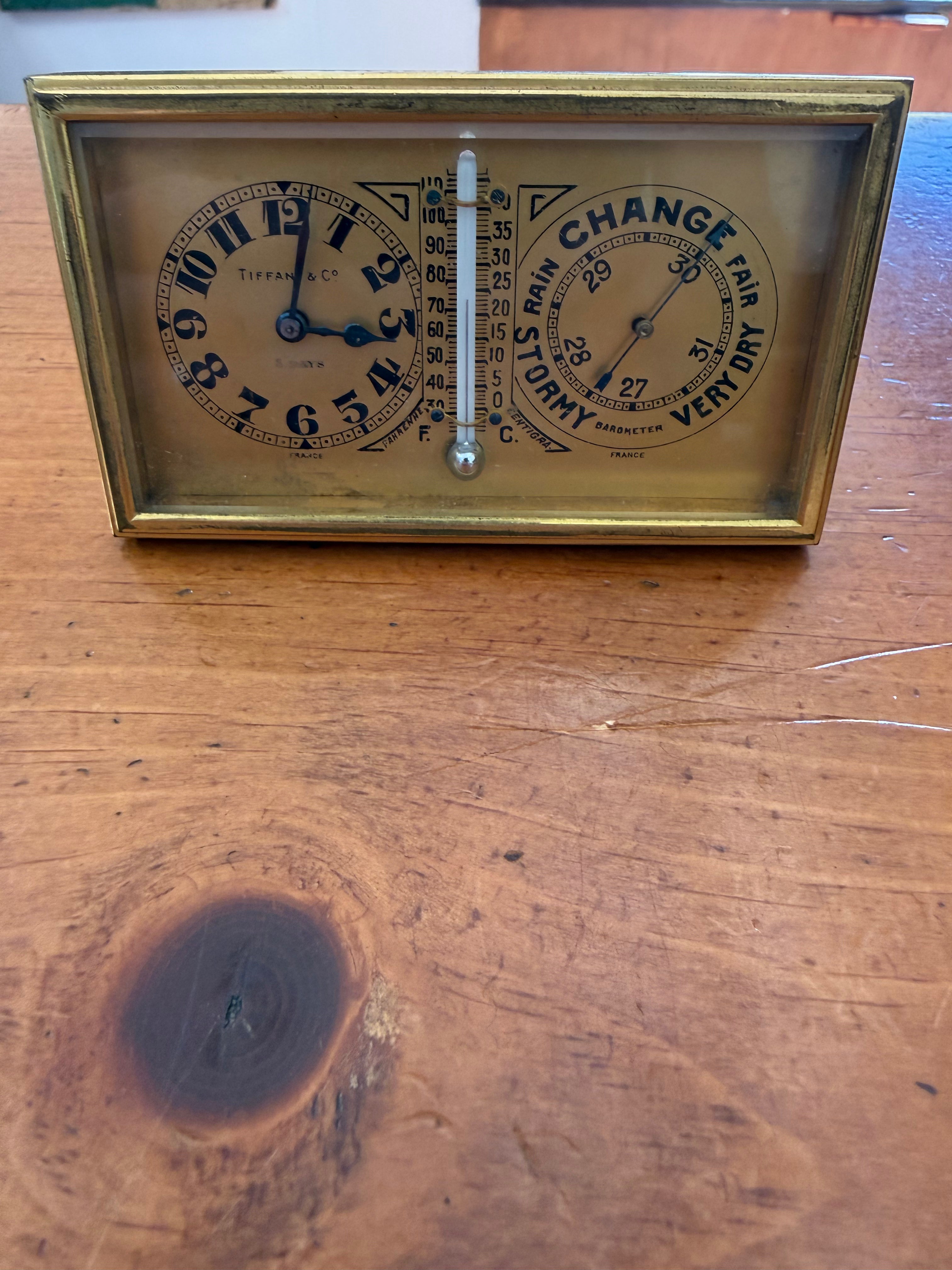 Front view of an antique Tiffany and Co. solid brass desk weather station showing a vertical rectangular layout with an 8-day clock dial at the top, a liquid thermometer measuring Fahrenheit and Centigrade in the middle, and a round aneroid barometer at the bottom marked Stormy, Rain, Change, Fair, and Very Dry