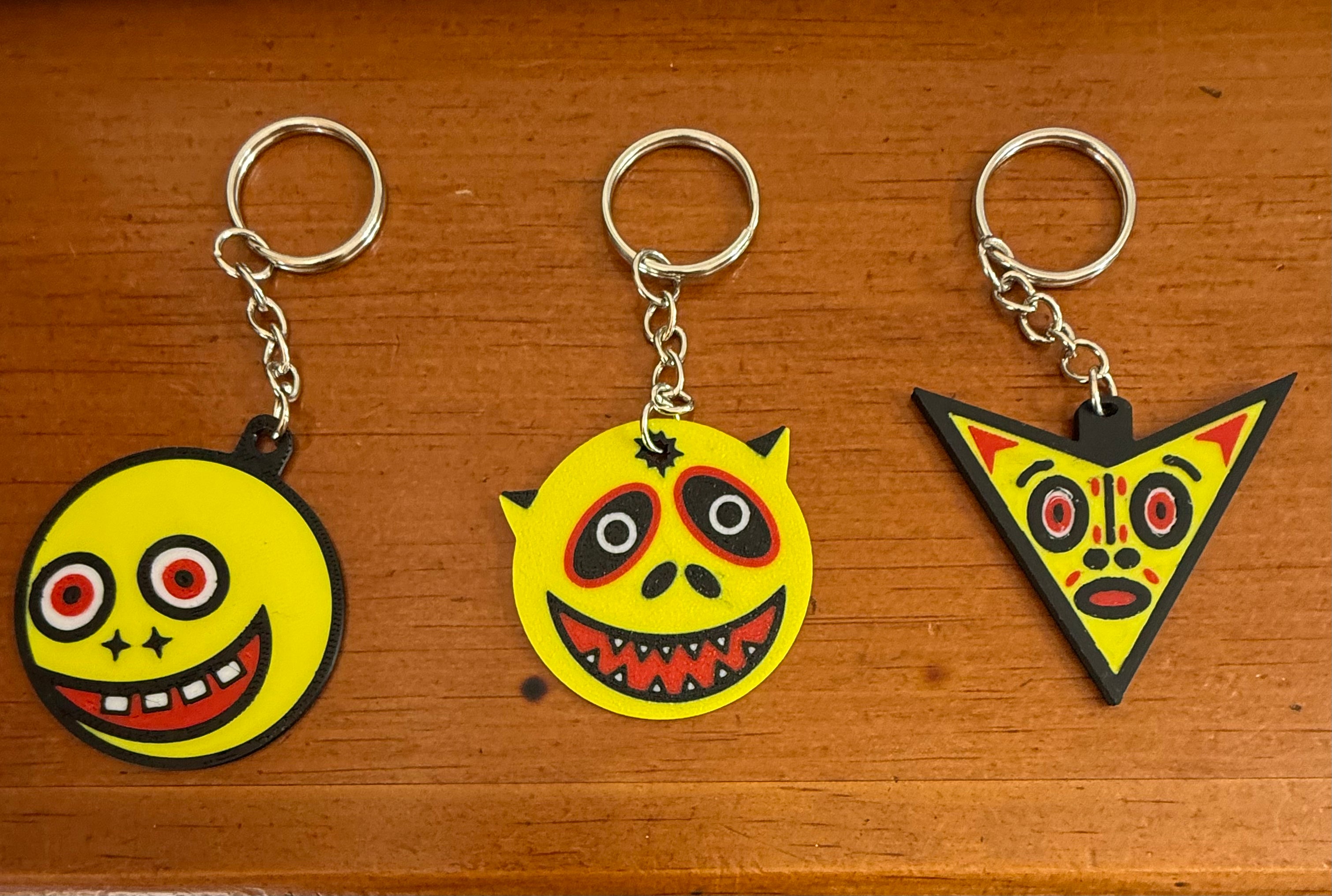 A trio of three unique lowbrow art keychains by Shelf4Life arranged on a wooden surface, each featuring a stylized yellow face with black outlines and red accents, including a round star-eyed smiling face, a spiked devil face with sharp teeth, and a downward triangle face with concentric eyes.