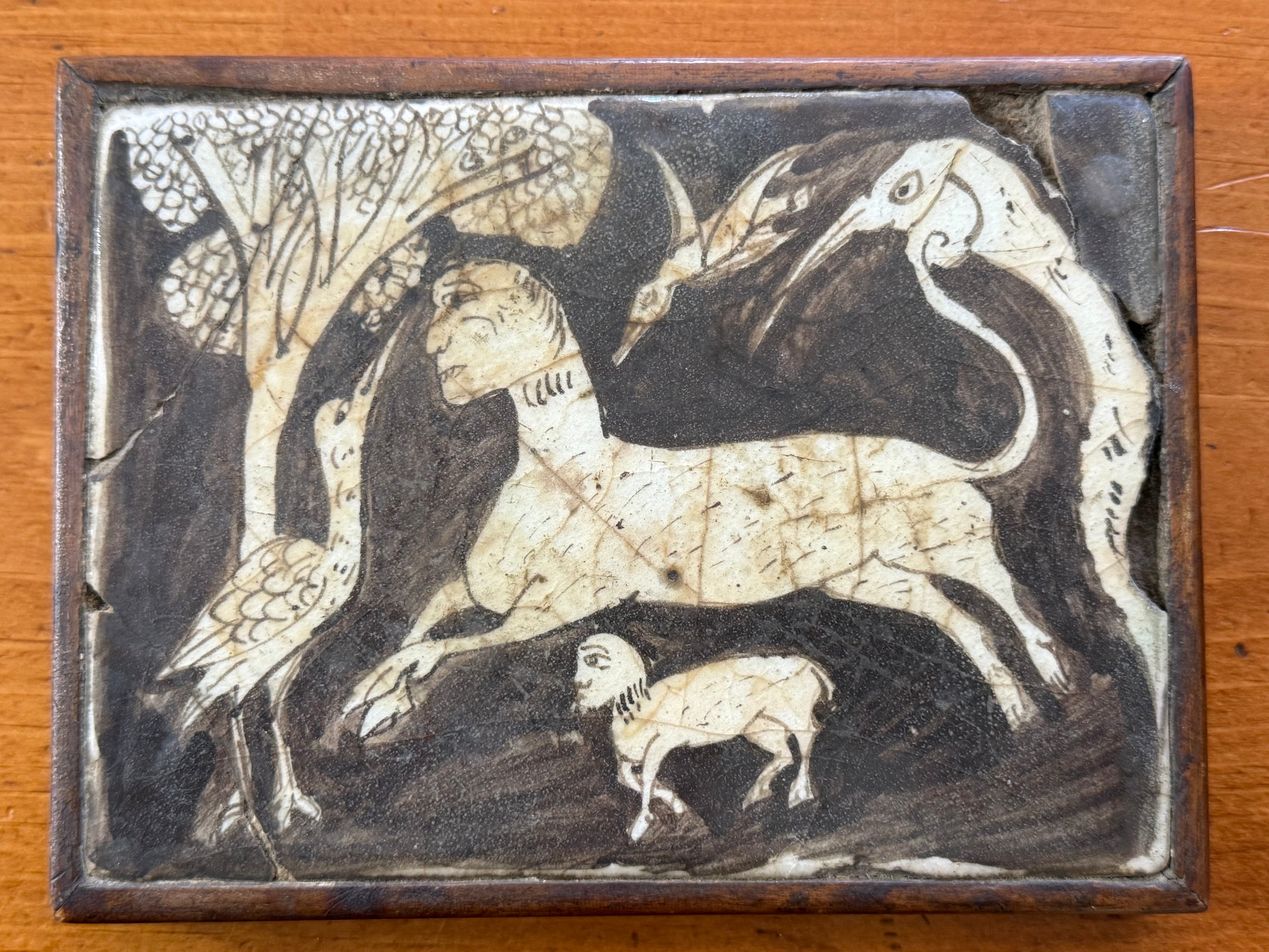A rectangular 10th to 12th century Persian sgraffito ceramic tile fragment featuring an incised cream-colored rearing lion or mythical beast against a dark slip background, surrounded by stylized birds and a textured tree canopy, showcased in a simple wooden frame.