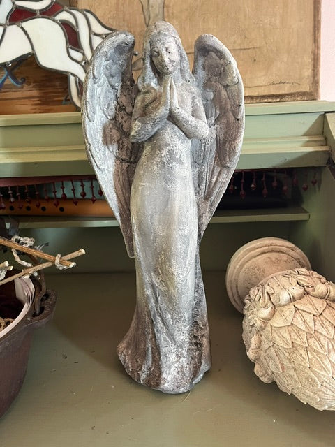 A vintage-style weathered concrete angel statue standing upright on a shelf, depicted with hands clasped in prayer and large wings spread wide, featuring a heavily distressed gray and brown textured finish.