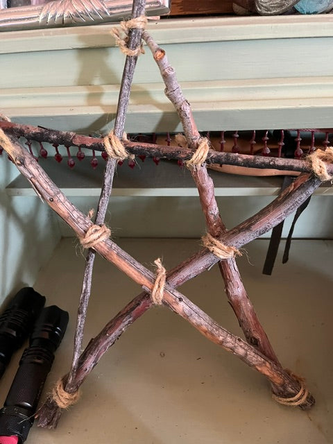 A handmade, five-pointed pentacle star constructed from rough-barked tree branches and securely bound at all joints with natural, textured jute twine, resting on a shelf. The rustic occult object, seen in Screenshot 2026-06-30 at 11.03.18 AM.png, has a raw, natural finish.