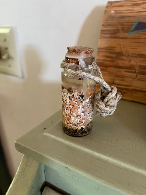 The vial is filled with distinctly layered ritual ingredients, including dark earth at the bottom and a thick middle layer of crushed white and brown eggshells. It is topped with a cork stopper sealed with drips of white wax, and a thick, knotted jute rope is tied securely around the bottle's neck.