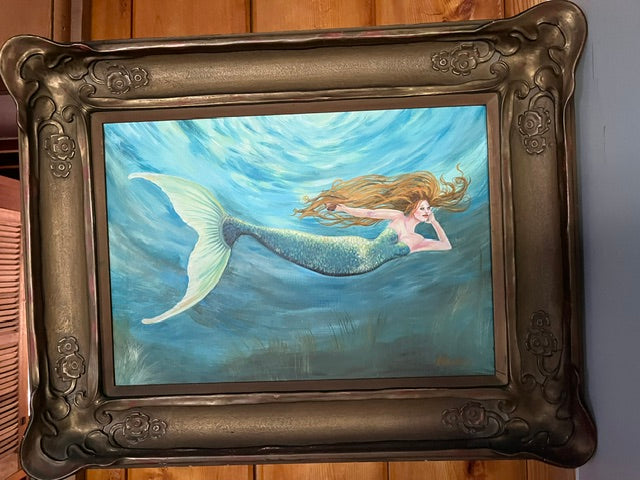 An original framed painting, depicting a mermaid with long red hair and a shimmering scaly tail swimming horizontally underwater through bright blue and turquoise sunlit waves. The artwork is set within a wide, heavy, and ornate dark metallic frame featuring embossed scroll and flower motifs.