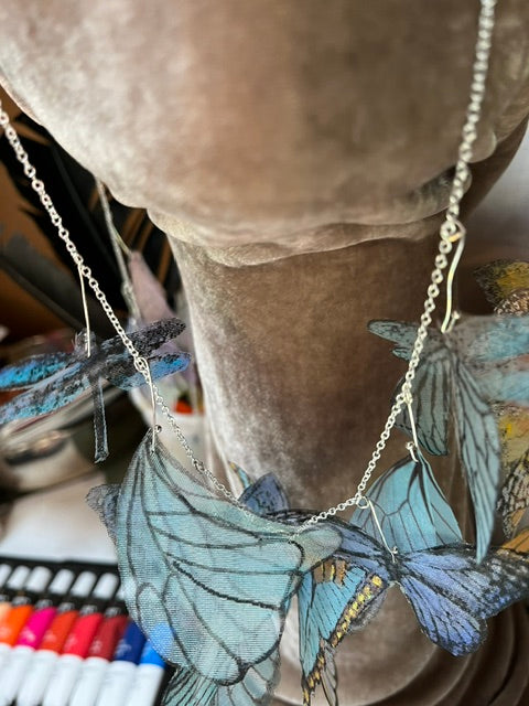 a unique statement necklace, featuring a delicate silver-tone metal chain adorned with multiple layered, translucent charms shaped like blue butterfly and dragonfly wings. The realistic printed wings display shades of light blue, teal, and navy with black vein patterns, hanging elegantly from a display bust.