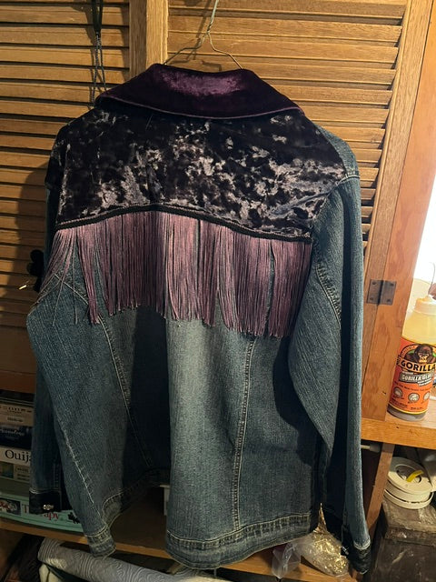 A medium-wash blue denim western jacket, The back of the jacket features a prominent upper yoke panel made of textured, dark purple crushed velvet, a smooth purple collar, and a thick row of long, lavender-colored fringe hanging horizontally across the shoulders.
