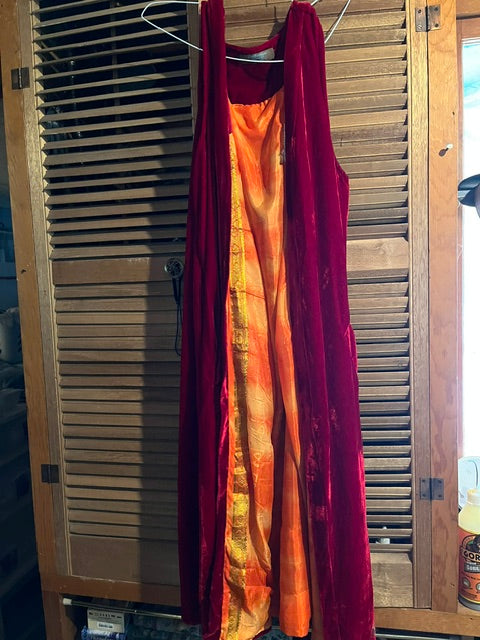 A longline, sleeveless open-front duster The garment features a rich red velvet exterior and a contrasting, bright orange and yellow textured interior lining, shown hanging against a background of rustic wooden slatted closet doors.