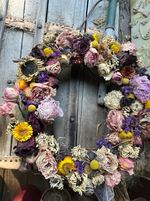 A rustic dried flower wreath made of pink, purple, yellow, and beige dried blooms, displayed on a weathered wooden surface.