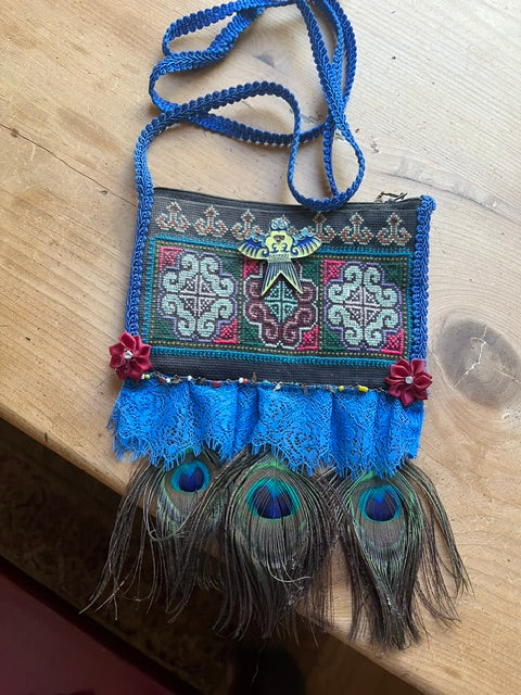A small handbag with a blue braided strap, blue lace trim, and three peacock feathers, featuring colorful embroidered geometric patterns on a dark background, resting on a light wood surface.
