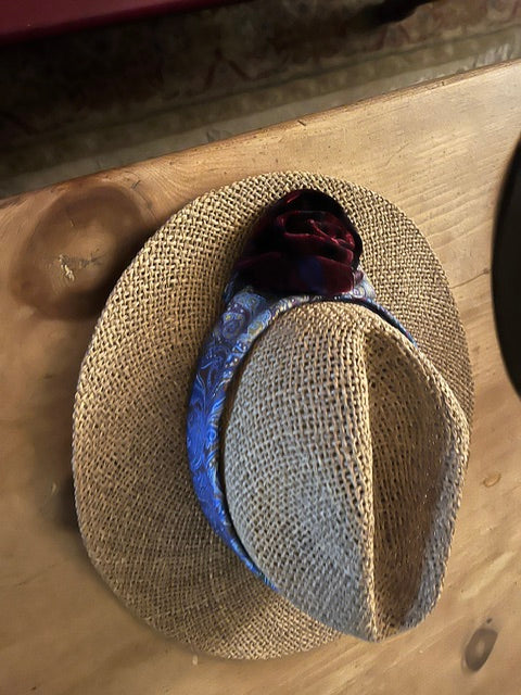 A woven straw fedora hat with a blue patterned band, a dark burgundy velvet rose, and a light beige textured band, displayed on a wooden table.