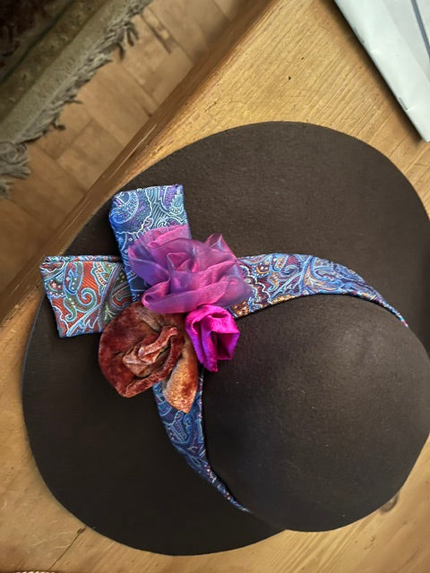 A dark brown felt hat with a blue paisley patterned ribbon band and decorative fabric flowers in purple, pink, and brown, resting on a wooden table.