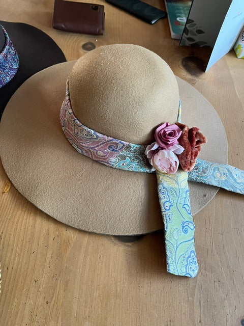 A beige wide-brim fedora hat with a paisley patterned headband, pink fabric roses, and a rust-brown scrunchie, displayed on a wooden table.
