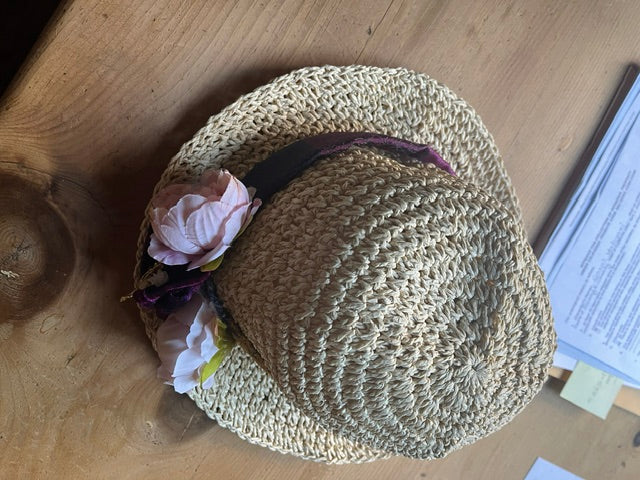 A rustic, woven beige straw sun hat decorated with a deep purple ribbon band and two light pink artificial peony flowers, resting on a natural wooden tabletop.