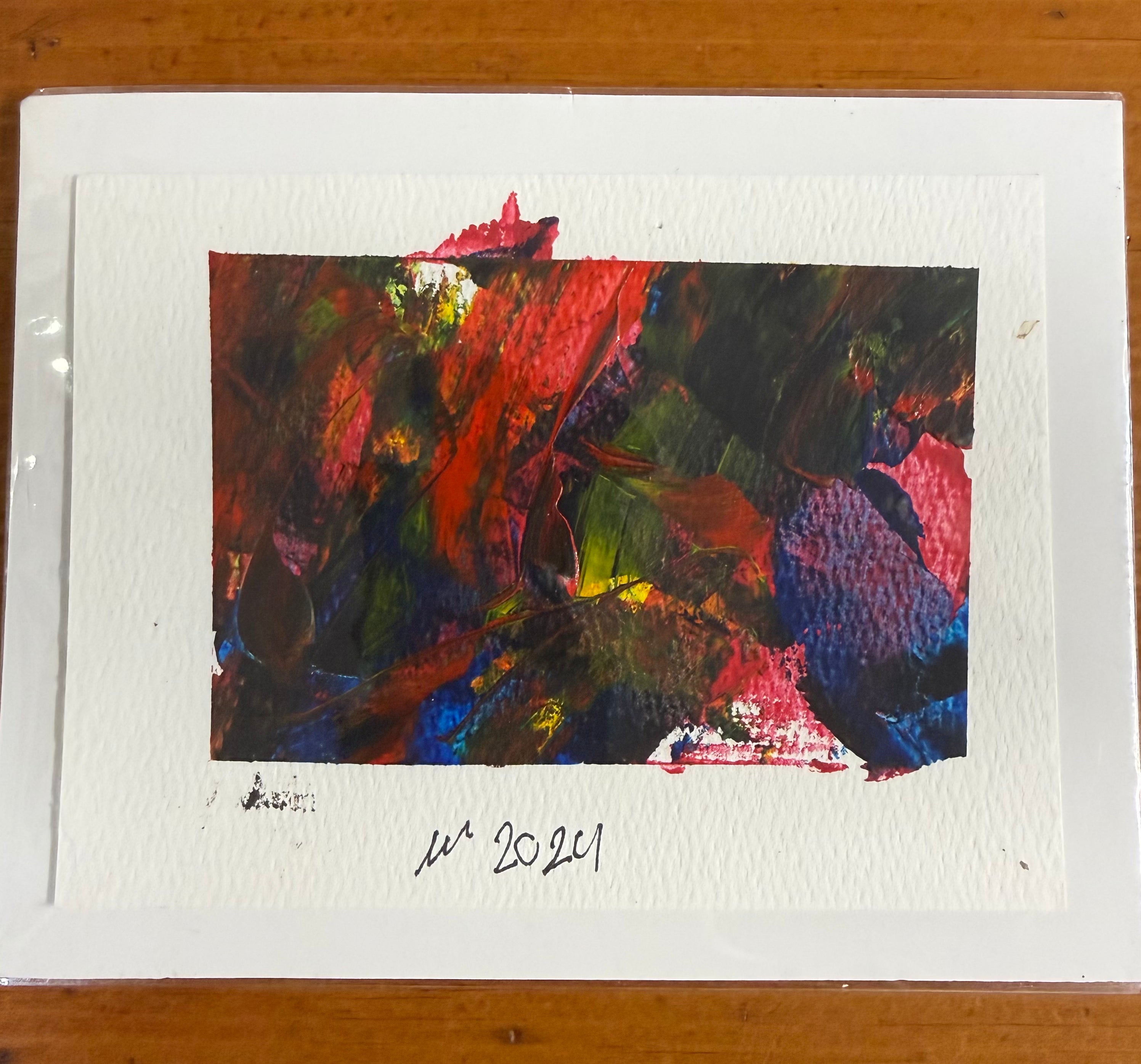 An abstract art print on textured white paper, featuring a colorful painted composition with red, green, blue, and black brushstrokes, placed on a wooden surface.