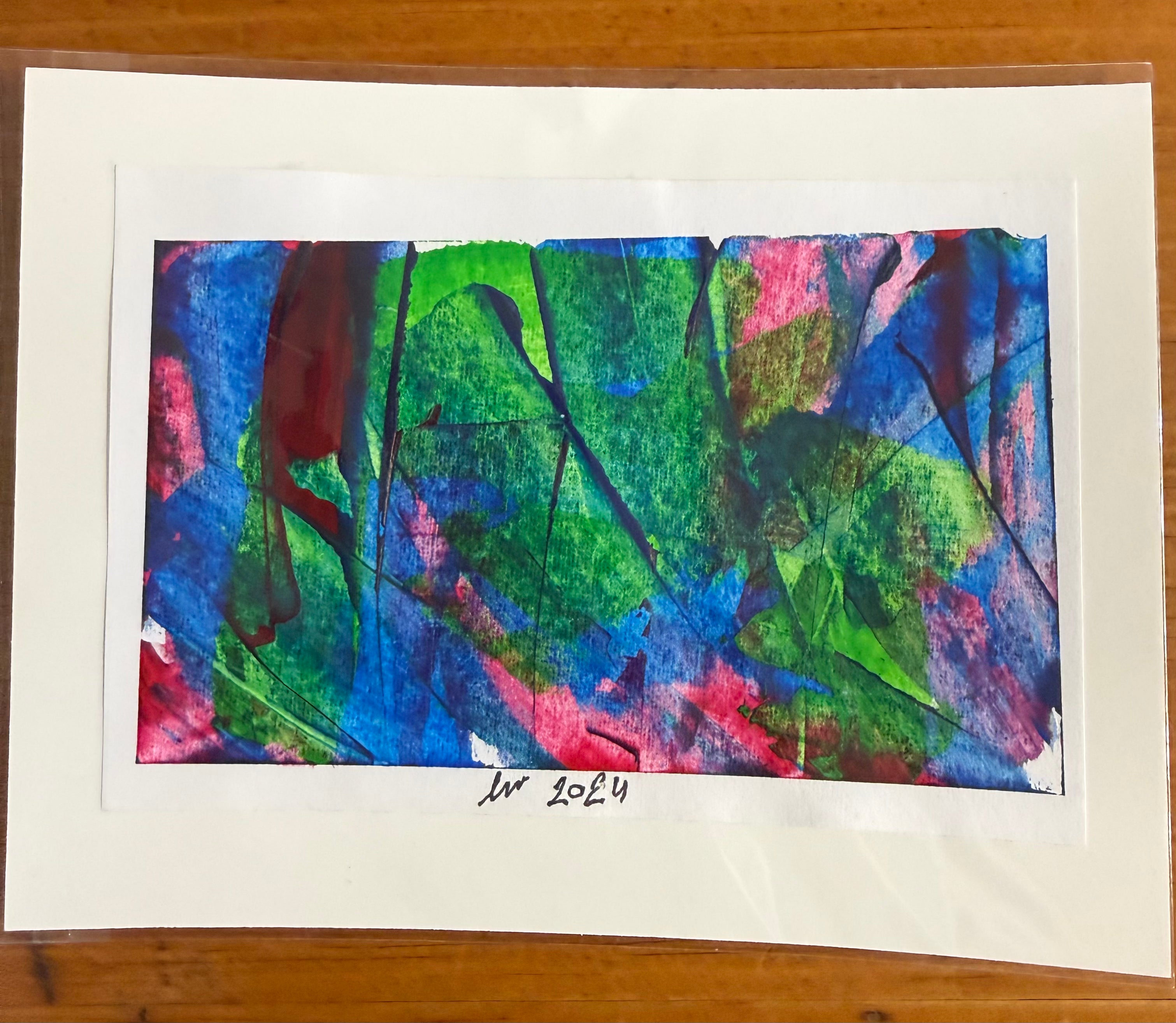 An abstract art print featuring vibrant green, blue, pink, and red brushstrokes, mounted on a wide white mat board with a handwritten signature and '1984' at the bottom, displayed on a wooden surface.