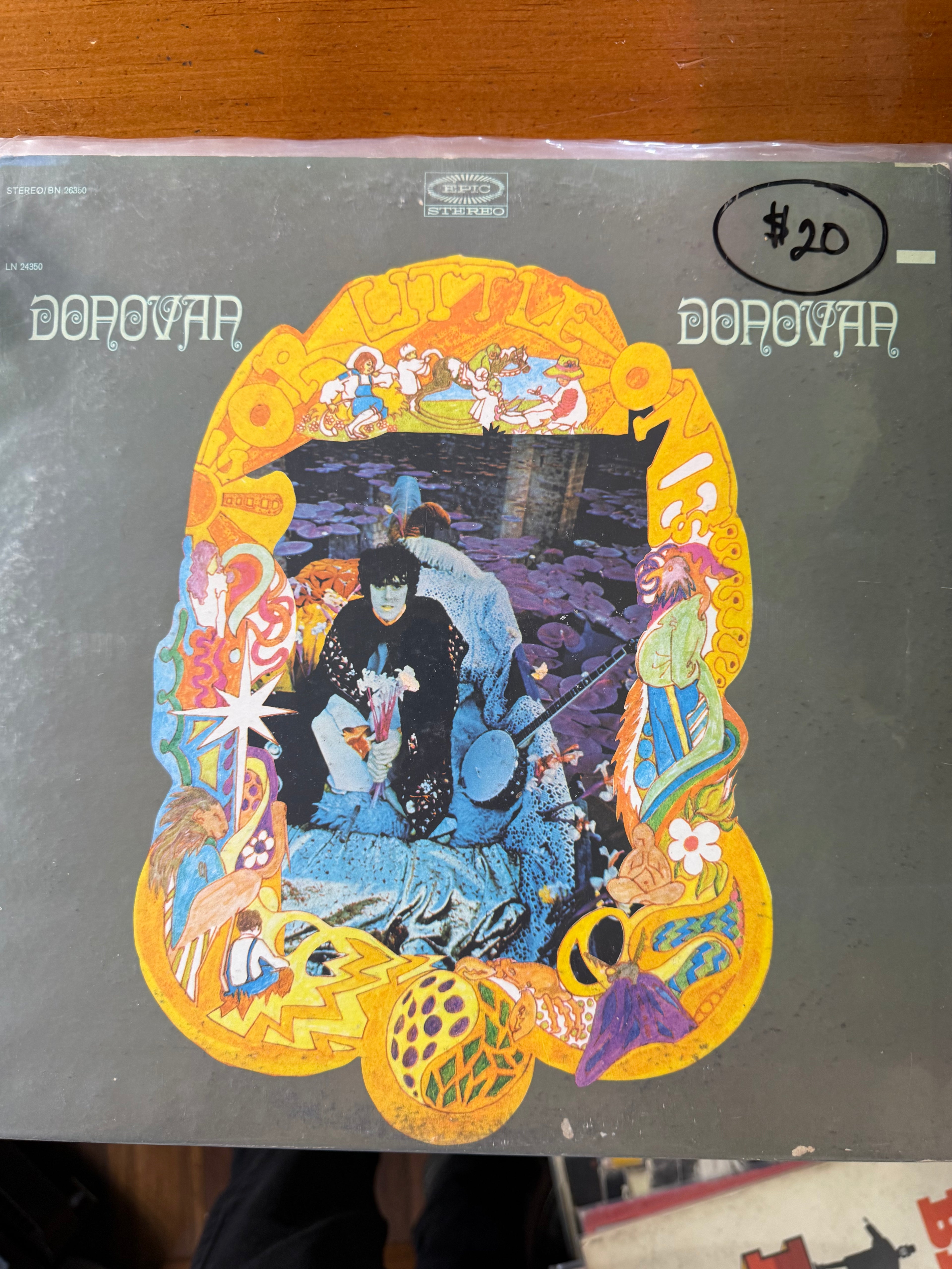 Front jacket cover of Donovan's For Little Ones vinyl album on Epic Records, featuring an ornate yellow psychedelic frame surrounding a photo of Donovan seated on a blue throne holding flowers next to a banjo.