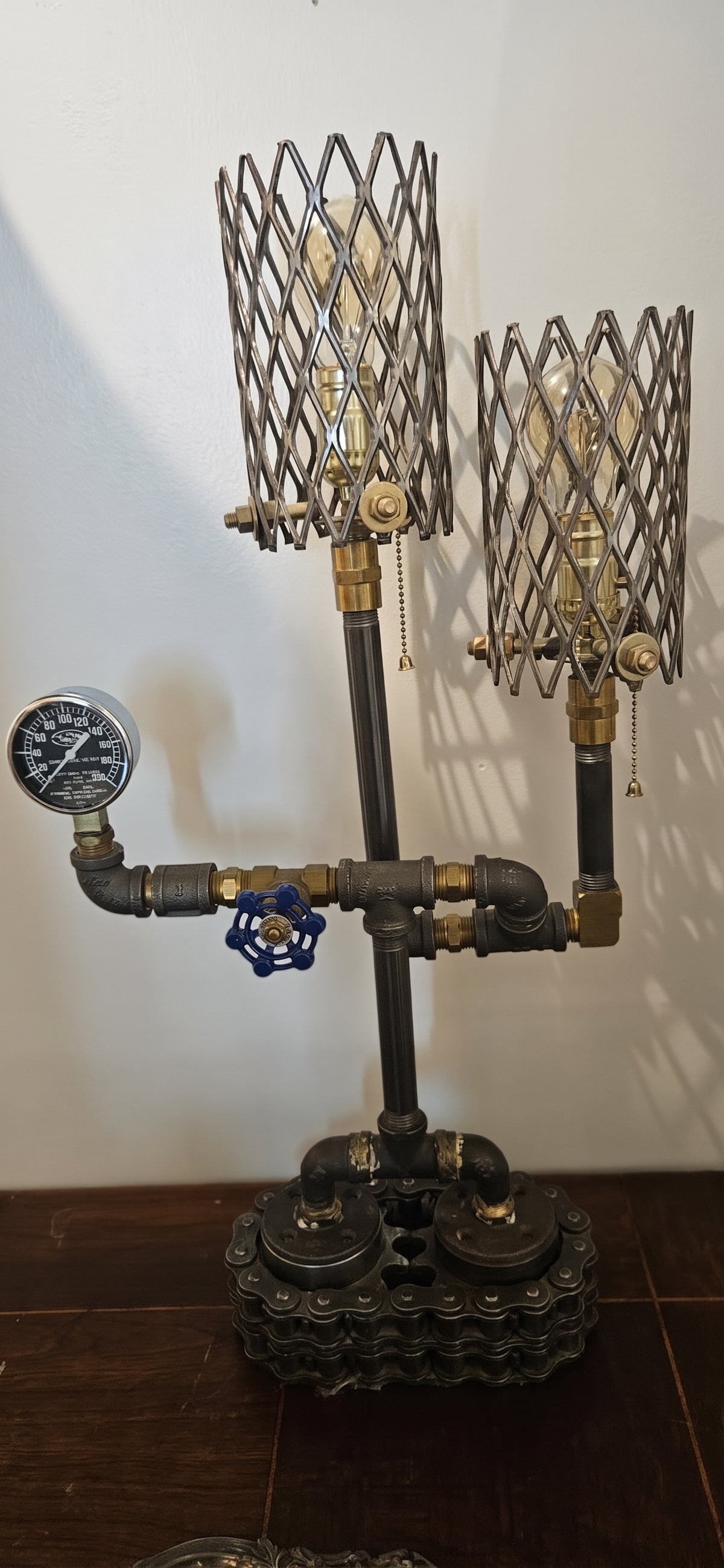 Handcrafted industrial steampunk pipe table lamp with two lights in wire mesh cage shades, an analog pressure gauge, and a heavy-duty chain-link metal base on a dark tabletop.