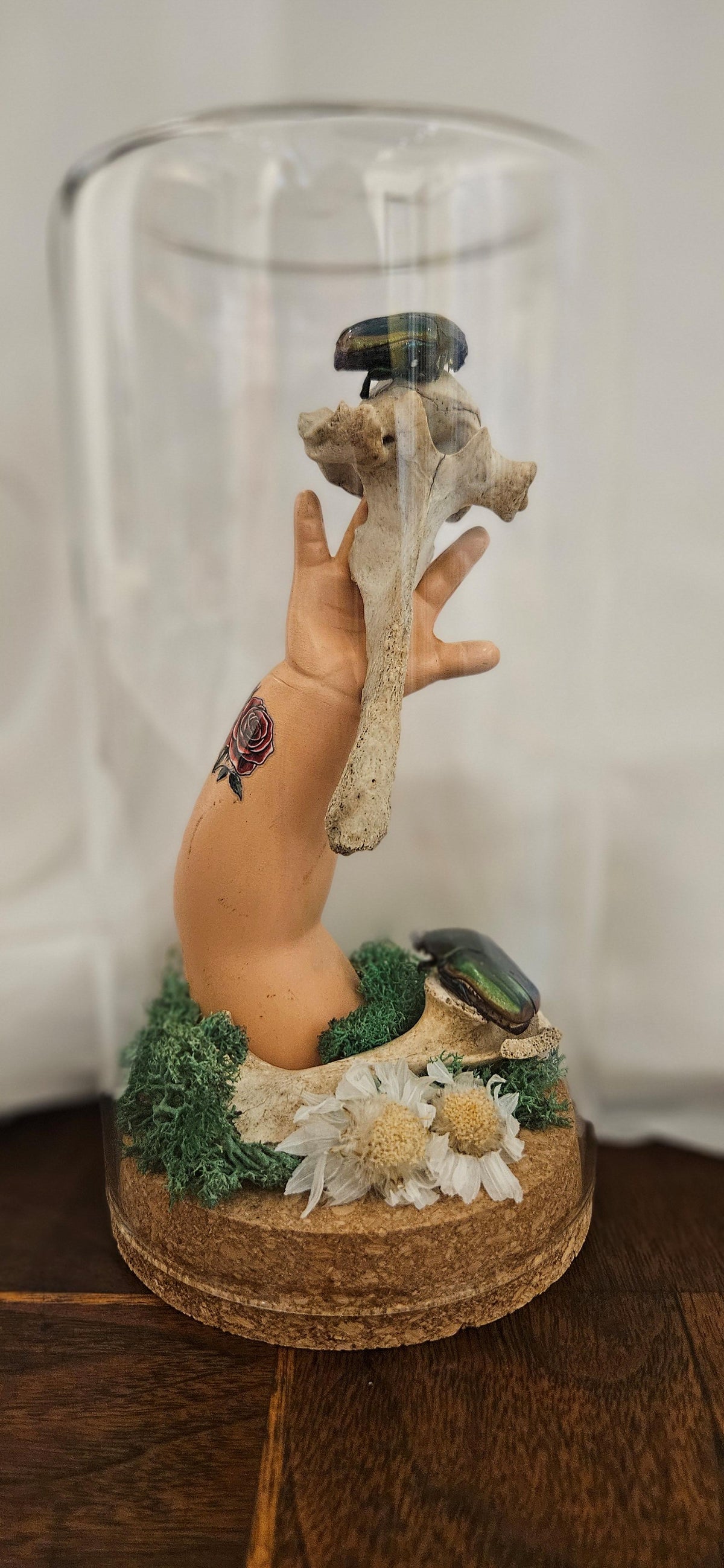 Handcrafted curiosity glass cloche featuring a tattooed doll hand reaching upward alongside an animal vertebrae bone, iridescent jewel beetles, and dried florals on a cork base.