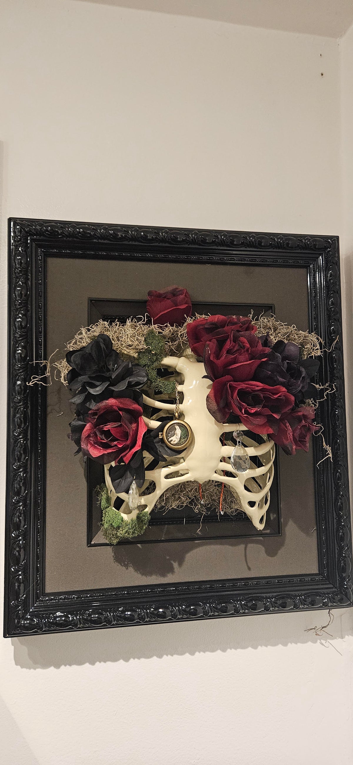 Ornate black frame displaying a faux rib cage adorned with dark red and black roses, Spanish moss, an antique cameo pendant, and dangling crystal prism drops.